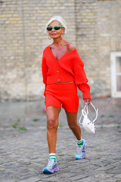 Grece Ghanem wears neon green sport sunglasses, gold with multicolored pearls pendant earrings, a neon red ribbed / wool / oversized sweater, a...