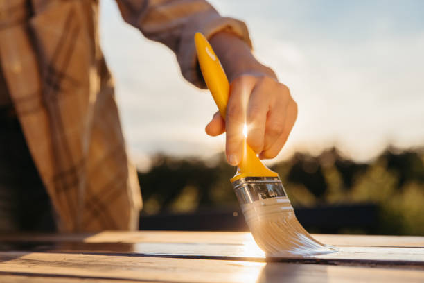 gros plan pinceau jaune tenu par une femme, table de peinture avec un rayon de soleil en vernis frais entre les doigts - garden decoration photos et images de collection