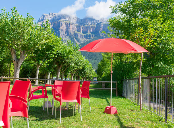 group of red chairs, tables and a parasol of a camping pub on green grass in torla - spain with the mountains of national park of ordesa at the background. horizontal photo. - garden decoration stock pictures, royalty-free ph