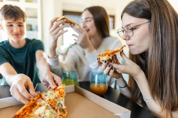 group of three teenagers eating pizza in a living room - junk food stock pictures, royalty-free photos & images