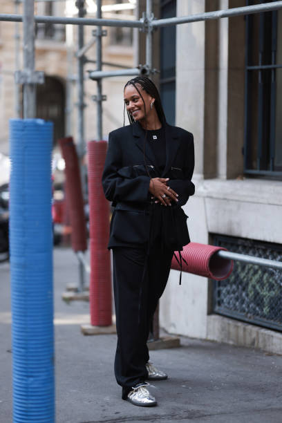 Guest is seen wearing black sunglasses, a black shiny suit with big attached bags and zippers on the blazer combined with black Botter shirt with...