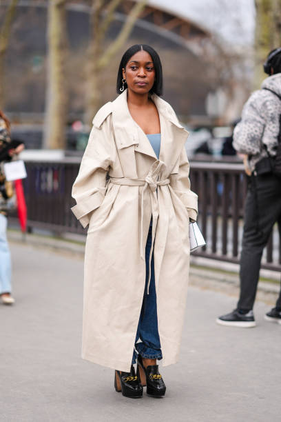 Guest wears a beige trench coat, a blue low neck top, a blue denim jeans / pants, black leather high heels shoes with golden chains, outside Chanel,...