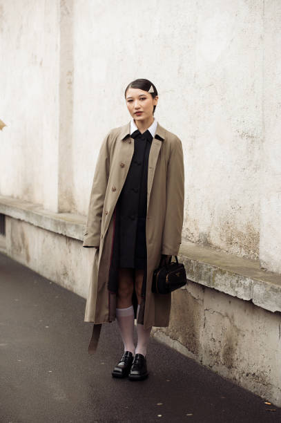 Guest wears a black dress, long white socks, black loafers, a brown trench coat, a black bag, and a metal Prada hair clip outside Prada during the...