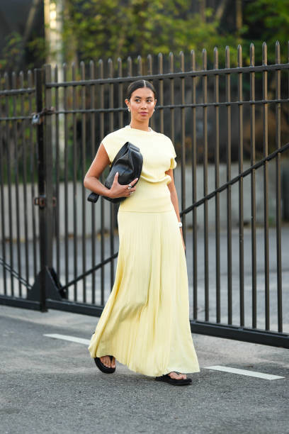 Guest wears a black leather bag, earrings, a yellow sleeveless top, a pale pastel yellow pleated long skirt, sandals, outside COS, during New York...