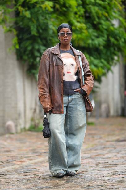 Guest wears a black silk scarf as a headband, black sunglasses, a black with beige face print pattern t-shirt, a dark brown shiny leather zipper...