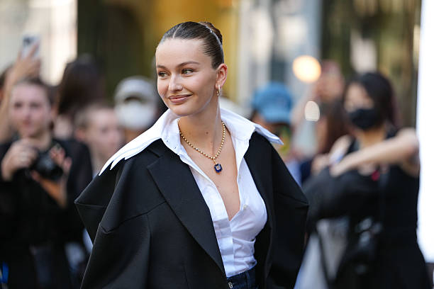 Guest wears a black structured blazer draped over the shoulders, paired with a crisp white button-up shirt featuring a wide collar. The hair is...