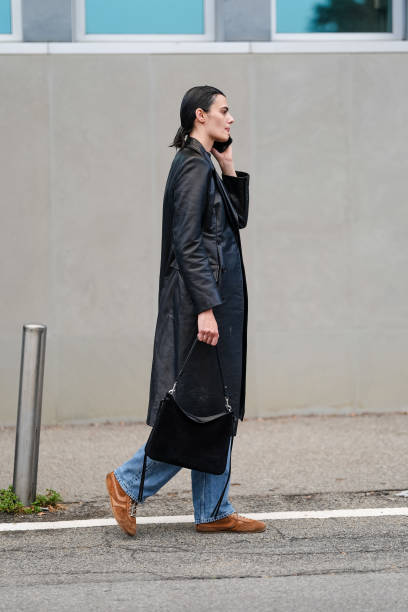 Guest wears a black t-shirt, a black leather trench coat, a black leather bag, blue denim jeans / pants, brown suede sneakers shoes, outside Jil...