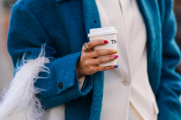 Guest wears a blue coat, a ring combined to a watch, holds a cup of coffee from Theory, has colored nail polish on the nails, outside Theory, during...