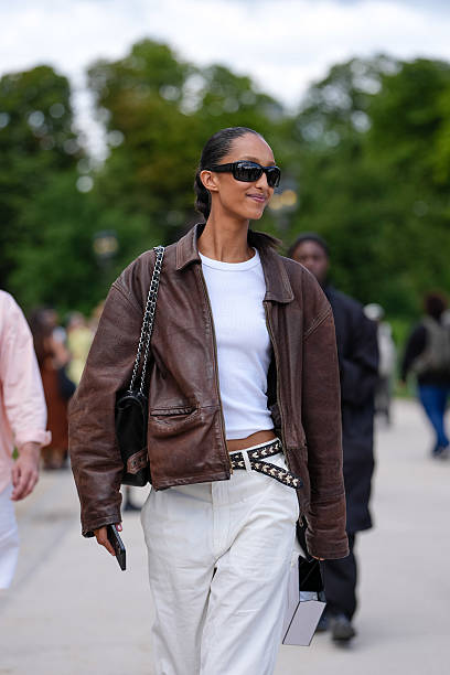 Guest wears a brown leather jacket with a classic collar and zip fastening. Underneath, a white fitted t-shirt is visible. The outfit includes white...