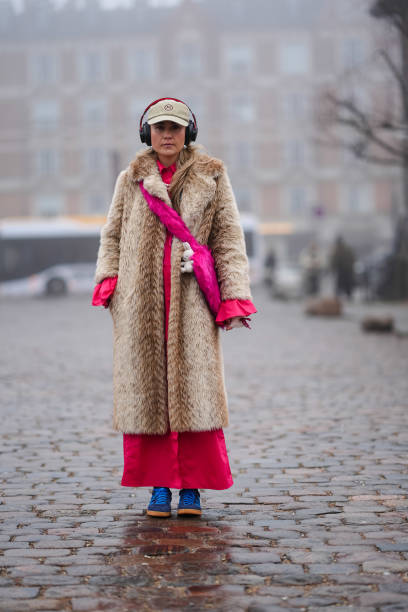 Guest wears a cap, a brown fluffy faux fur long coat, a pink dress, blue sneakers, headphones, outside Rolf Ekroth, during the Copenhagen Fashion...