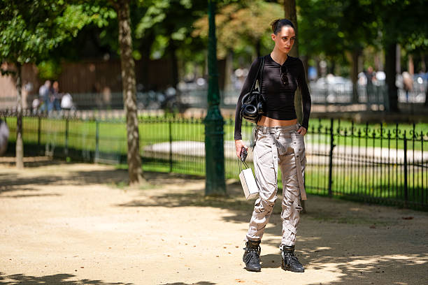 Guest wears a fitted, long-sleeve black top with a round neckline. Sunglasses are tucked into the neckline. Hair is styled in a sleek bun. Carries a...