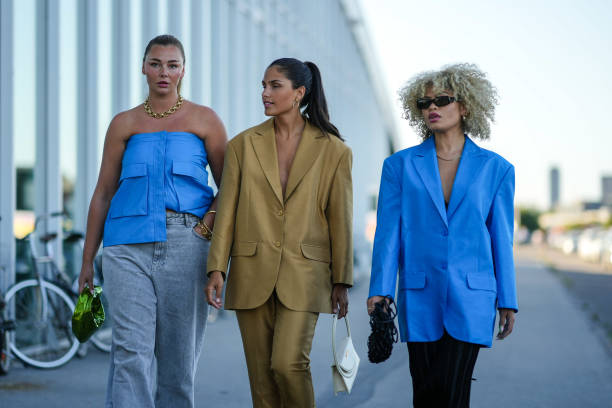 Guest wears a gold large chain necklace, a blue leather shoulder-off top, gray denim large pants, a green shiny leather handbag, a guest wears gold...