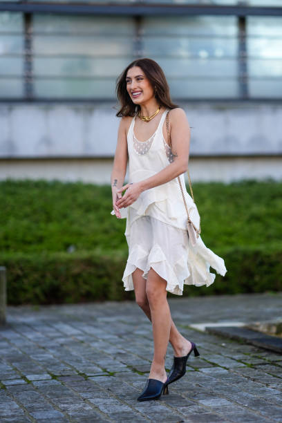 Guest wears a golden necklace, a white bag, a white sleeveless dress with a handkerchief lower part, black heeled mules, outside Stella McCartney ,...
