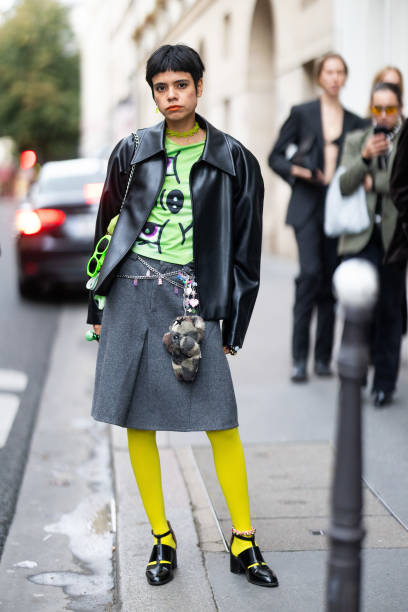 Guest wears a green necklace, a green printed t-shirt, a black leather jacket, a silver belt, a grey skirt, fluo green sunglasses, yellow tights and...