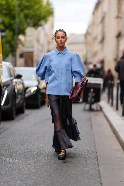Guest wears a light blue oversized shirt, a black transparent long skirt, a burgundy leather bag, black leather shoes, outside Sacai, during...