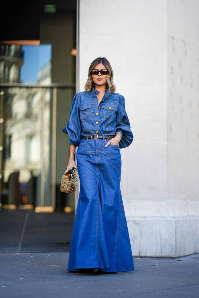 Guest wears a long blue denim dress, a black leather belt, a gold bag, sunglasses, outside Zimmermann, during the Womenswear Fall/Winter 2024/2025 as...