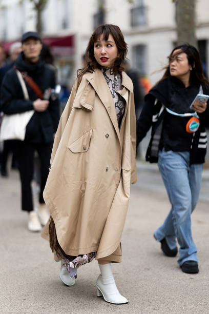 Guest wears a snake leather printed turtle neck shirt, a beige long trench and white leather heeled ankle boots outside Stella McCartney show during...