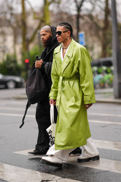 Guest wears a white jacket as a top, a green leather trench coat, white pants, white leather boots, a white tote-bag, sunglasses, outside Hermes,...