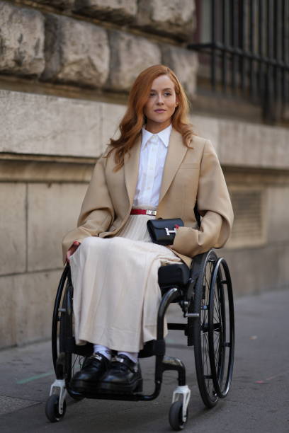 Guest wears a white shirt, a beige oversized blazer jacket, a belt, pleated skirt, a black mini Hermes bag, outside Hermes, during the Womenswear...