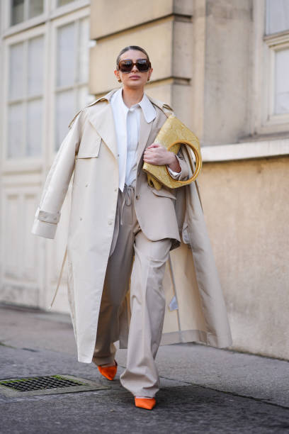Guest wears a white shirt, a beige trench coat, beige wide-leg pants, orange pointed shoes, a yellow leather bag, sunglasses, outside Giambattista...