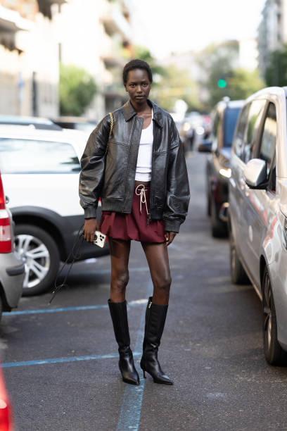 Guest wears a white tank top, a red burgundy mini puffy skirt, black leather boots and a brown leather jacket outside Max Mara show during the Milan...