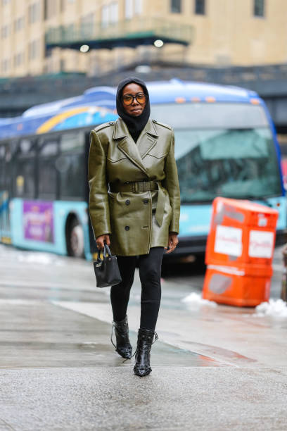 Guest wears black balaclava, black sunglasses with orange lenses, shiny olive green belted double breasted leather coat from Prada, black mini...