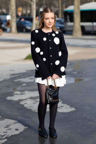 Guest wears black cardigan with white flowers decorations, white mini skirt, black tights, black Chanel bag, black Chanel mary jane shoes, outside...