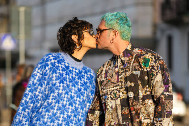 Guest wears black square sunglasses, silver earrings, a black high neck t-shirt, a blue and royal blue print pattern oversized pullover, a guest...