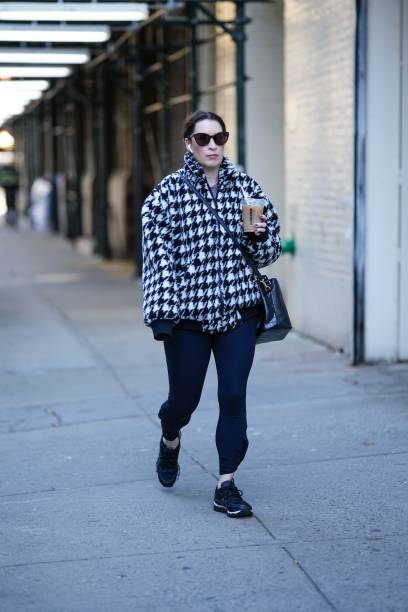 Guest wears black sunglasses, a black and white houndstooth print pattern oversized bomber coat, a black shiny leather crossbody bag, black legging...