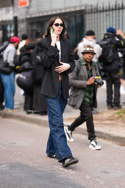 Guest wears black sunglasses, a black blazer jacket, a white top, a black leather bag, blue denim jeans / pants, black and white sneakers shoes,...