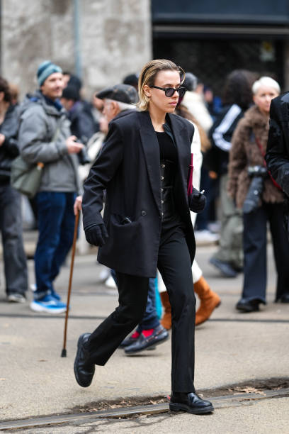Guest wears black sunglasses, a black oversized blazer jacket, black pants, black gloves, black leather shoes, outside Dolce & Gabbana, during the...