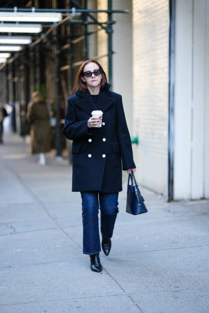 Guest wears black sunglasses, a black t-shirt, a black buttoned coat, a navy blue shiny leather handbag, navy blue pants, black shiny leather pointed...
