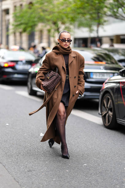 Guest wears black sunglasses, a brown scarf, a brown oversized coat, a dark brown top, a black mini skirt, black tights, brown leather bag, black...