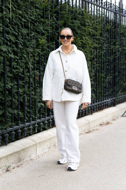 Guest wears black sunglasses, a Fendi cross-body Baguette bag, a gold necklace, white sneakers, a white jacket and white flared trousers outside...