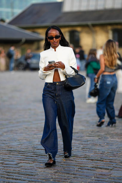 Guest wears black sunglasses, a white high neck / jacket, a black shiny leather handbag, navy blue denim wide legs pants, black velvet ballerinas ,...