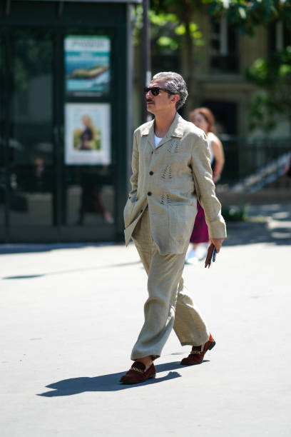 Guest wears black sunglasses, a white t-shirt, a beige buttoned with cut-out leaves pattern jacket, matching beige linen large pants, orange camel...