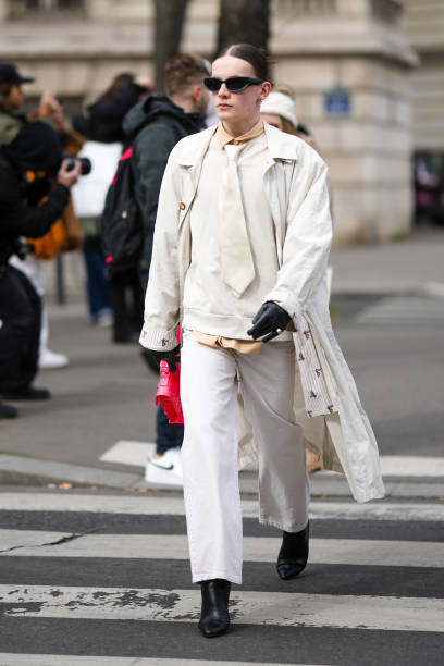 Guest wears black sunglasses, a white tie, a white latte sweater, a white long belted coat, white denim large pants, black shiny leather gloves, a...