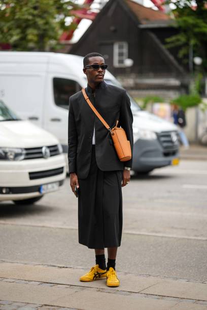 Guest wears black sunglasses, an orange shiny leather crossbody bag, a black blazer jacket, a white t-shirt, a black midi skirt, black socks, yellow...
