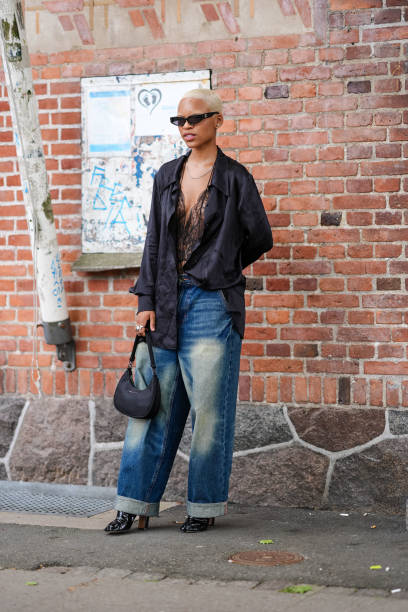 Guest wears black sunglasses, black lace sheer mesh bodysuit, shiny black oversized long sleeve shirt, silver rings, washed navy blue denim jean...