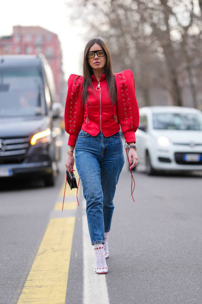 Guest wears black sunglasses, bright cherry red puffy sleeve zipped jacket, navy blue form fitting denim jean pants, gold bracelets, a black gold...