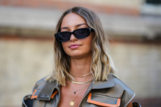 Guest wears black sunglasses, gold chain pendant necklaces, a dark gold shiny sleeveless / zipper pockets / cargo oversized jacket, during the...