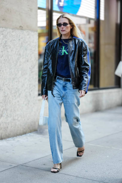 Guest wears black sunglasses, gold necklace, dark navy blue bright green Calvin Klien Jeans print t-shirt, shiny black dark blue lined unzipped biker...
