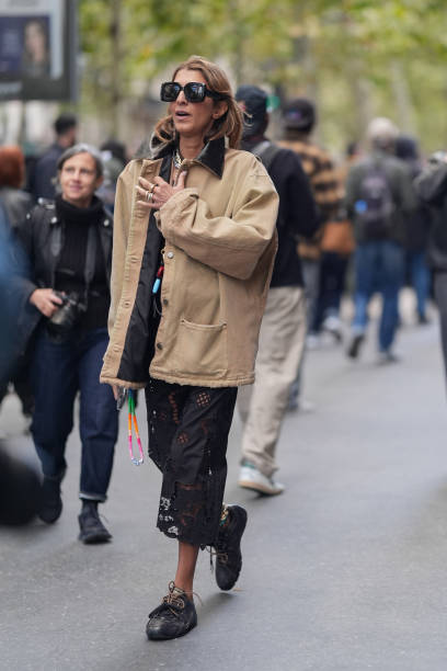 Guest wears black sunglasses, light brown / cream jacket, a black lace skirt with floral details, black sneakers shoes, outside Hermes, during...