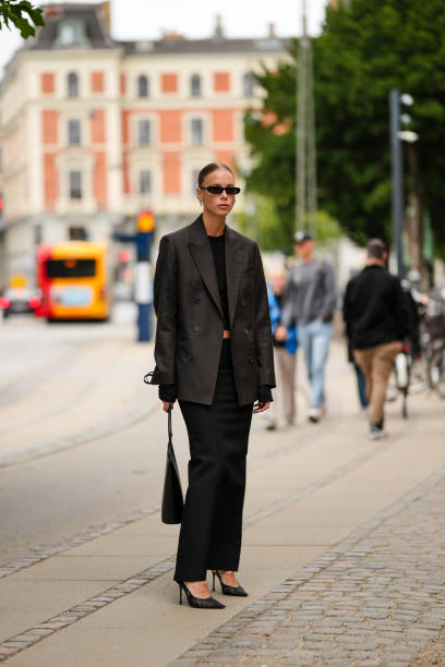 Guest wears black sunglasses, silver earrings, a black cropped top, a black oversized blazer jacket, a black high waist long skirt, a black shiny...
