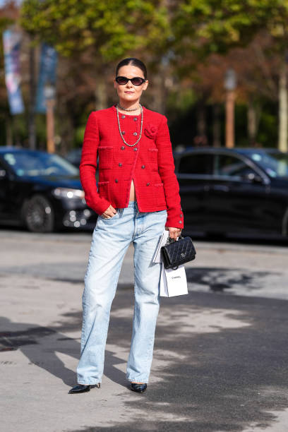 Guest wears black sunglasses, silver layered necklaces, bright red tweed Chanel blazer jacket, washed light blue denim jean pants, shiny black...