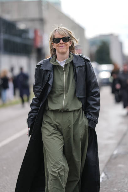 Guest wears black sunglasses, white t-shirt, dark olive green zipped bomber jacket, dark olive green loose trouser pants, shiny black leather trench...