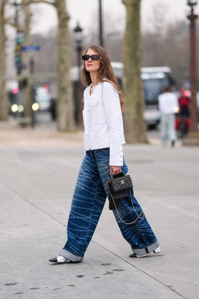 Guest wears black sunglasses, white tweed pearl detail buttoned Chanel blazer jacket, navy blue white pearl pattern/print baggy Chanel denim jean...