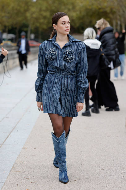 Guest wears blue denim striped shirt dress with flowers detail, black sheer tights, denim cowboy boots, outside Balenciaga, during Womenswear...