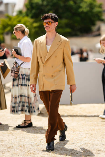 Guest wears brown and orange sunglasses, a white t-shirt, a yellow buttoned long blazer jacket, dark brown velvet suit pants, black shiny leather...