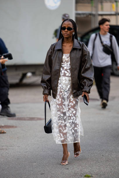 Guest wears brown leather jacket, white transparent dress outside Baum & Pferdgarten during day three of the Copenhagen Fashion Week SS25 on August...
